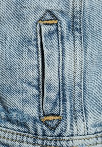 Světle modrá denimová tkanina s kapsou ve tvaru diamantu a žlutým prošíváním. Materiál vykazuje mírné třepení a variace textury.