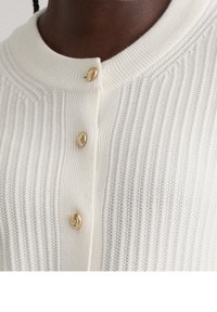 Bílý žebrovaný pletený kardigan s kulatým výstřihem a třemi zlatými knoflíky. Textura má vertikální pruhy a jemný povrch.