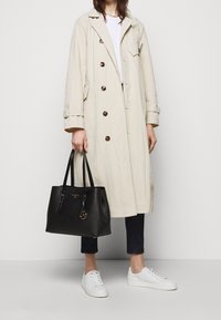 Beige trench coat dengan tombol coklat, tas tote kulit hitam dengan aksen emas, jeans gelap, dan sneakers putih. Desain yang sederhana dan fungsional.