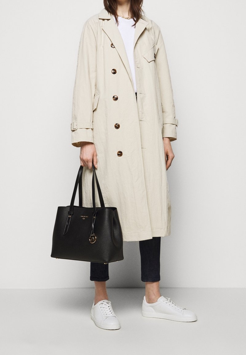 Beige trench coat dengan tombol coklat, tas tote kulit hitam dengan aksen emas, jeans gelap, dan sneakers putih. Desain yang sederhana dan fungsional.