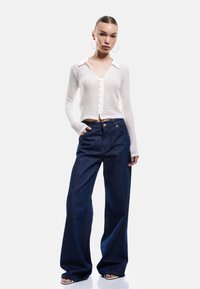 Weißer, strukturierter Cardigan mit langen Ärmeln und V-Ausschnitt, der mit hoch taillierten, weit geschnittenen, dunkelblauen Jeans kombiniert wird.