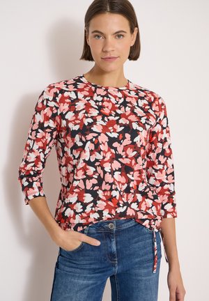 Femme portant un haut à imprimé floral rouge, rose, blanc et noir avec des manches trois-quarts et un jean bleu, debout devant un fond uni.