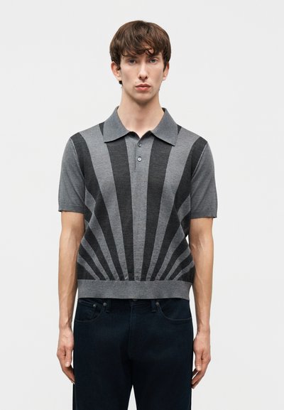 Mann trägt ein graues Poloshirt mit kurzen Ärmeln und schwarzen radialen Streifen, kombiniert mit dunklen Jeans, steht vor einem schlichten Hintergrund.