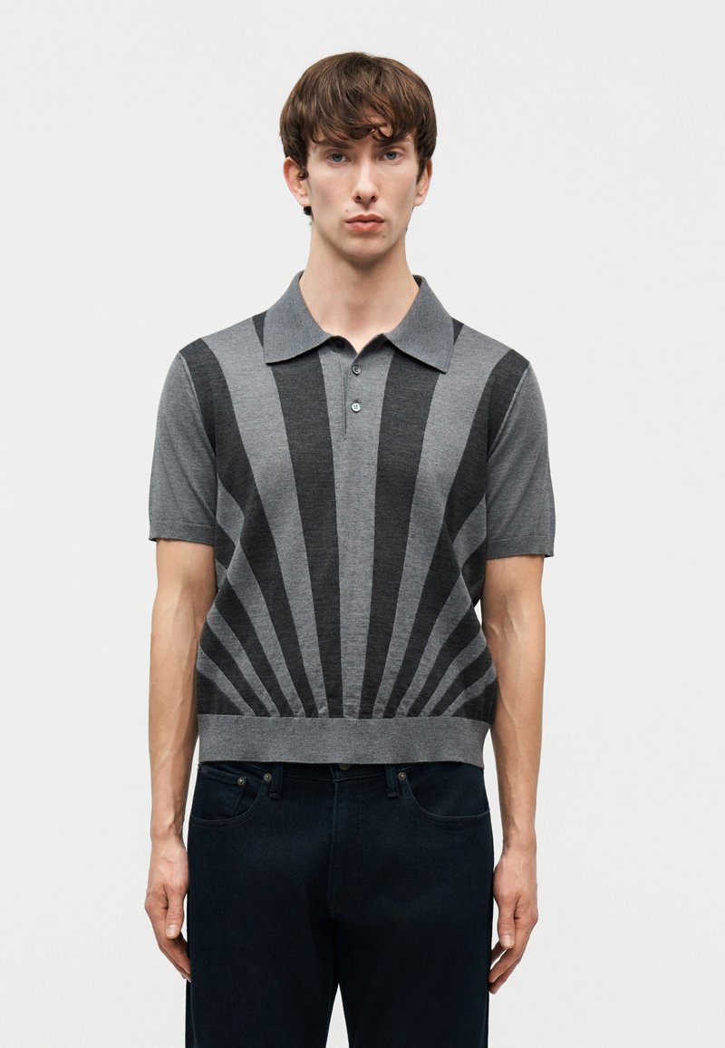 Άνδρας που φοράει γκρι πόλο με κοντά μανίκια και μαύρες ακτινωτές ρίγες, συνδυασμένος με σκούρα τζιν, στέκεται μπροστά σε απλό φόντο.