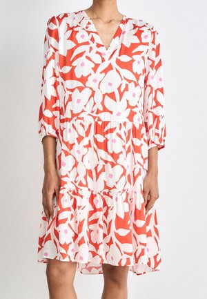 Kvinde iført knælængde hvid kjole med lys rød og pink blomsterprint, trekvartærmer og V-udskæring.