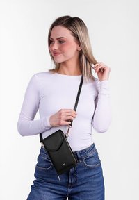 Černá kabelka přes rameno se zipovým uzávěrem, hladkou koženou texturou, obdélníkovým tvarem a odnímatelným popruhem, kterou drží osoba v světlém topu.