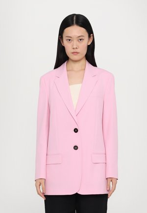 Blazer rosa con revers a punta, due tasche frontali e due bottoni neri. Texture liscia, vestibilità su misura, abbinato a una maglietta chiara e pantaloni scuri.
