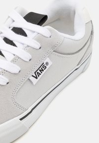 Tenisi gri din pânză cu șireturi albe, având un tab de Accentuare negru și logo-ul Vans. Talpa din cauciuc cu detalii cu dungi negre pe margine.