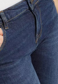 Ciemnoniebieskie dżinsy o dopasowanym kroju, z kontrastowymi żółtymi szwami i metalowym guzikiem w pasie.