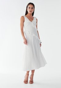 Weißes Midi-Kleid mit rüschiertem Ausschnitt und Saum, aus strukturiertem Stoff und mit tailliertem Oberteil. Akzentuiert mit dünnen Trägern und dezenten Details.