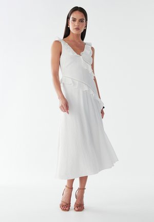 Robe midi blanche avec un col et un ourlet à volants, mettant en avant un tissu texturé et un corsage ajusté. Accentuée par des bretelles fines et des détails subtils.