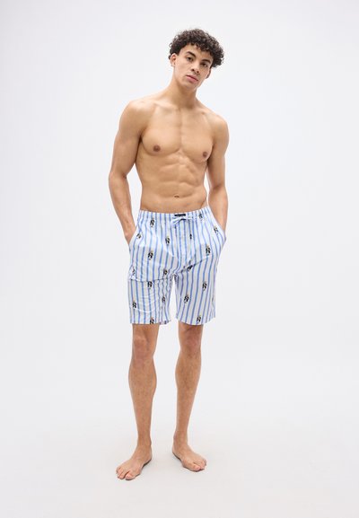 Άνδρας χωρίς πουκάμισο, όρθιος ξυπόλυτος, φοράει λευκό και μπλε ριγέ σορτς με μικρά σχέδια αρκουδάκι και έχει τα χέρια στις τσέπες.