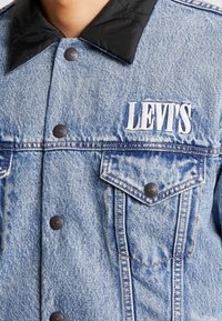 Ljusblå denimjacka med svart vadderad krage, förladdad med framfickor, knäppning med knappar och vit "LEVI'S" logotyp på bröstet.