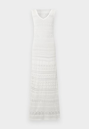 Λεπτή γυναίκα που φοράει λευκό αμάνικο πλεκτό μακρύ φόρεμα και κίτρινα σανδάλια, στέκεται μπροστά σε ένα απλό λευκό φόντο.