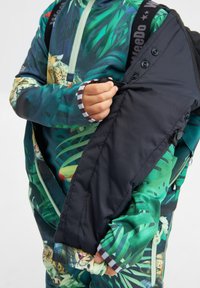 Giacca colorata con un design a foglie e maculato, caratterizzata da una manica nera imbottita e dettagli a righe sui polsini. Chiusura con zip e cintura regolabile.