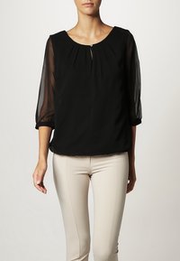 Femme portant un chemisier noir à manches trois-quarts transparentes et un pantalon beige ajusté, debout devant un fond clair uni.