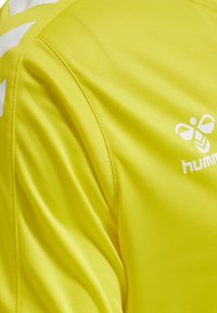 Gele sporttrui van glad, ademend materiaal. Bevat een wit logo en accentstrepen langs de schouders. Close-up weergave.