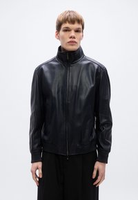 Fekete bőrkabát magas gallérral, cipzáras lezárással és bordázott mandzsettákkal. Simas textúrájú, oldalzsebekkel és testhezálló szabással tervezve.