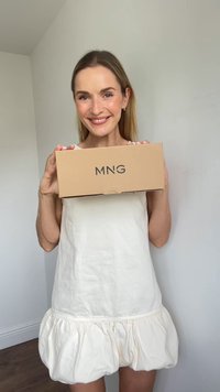 Donna sorridente in abito bianco che tiene in mano una scatola marrone con il logo "MNG", in piedi contro un muro chiaro e semplice all'interno.