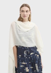 Écharpe blanche légère et semi-transparente drapée sur les épaules, avec une texture douce et un accent vert brodé sur un bord.