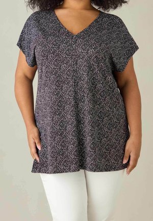 Vrouw draagt een zwart-wit patroon korte mouwen V-hals top met een witte broek tegen een effen achtergrond.