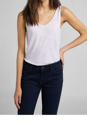 Kobieta ubrana w biały, bezrękawnikowy top z dekoltem w serek i ciemnoniebieskie jeansy, z jedną ręką na biodrze, a drugą swobodnie przy boku.