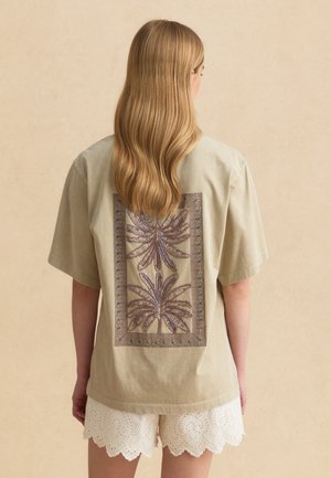 Donna con capelli biondi lunghi che indossa una maglietta beige oversize con motivo di foglia di palma ricamato sulla schiena e pantaloncini bianchi a intaglio.