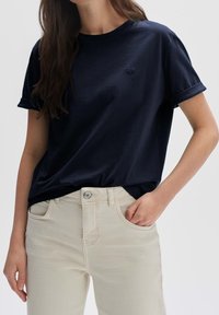 Maglietta blu navy a maniche corte in tessuto morbido, con un logo a cuore discreto sul petto sinistro. Abbinata a jeans di colore chiaro.