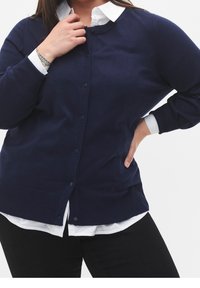 Žena v námořnickém modrém cardiganovém svetru s knoflíky přes bílou košili a černé kalhoty, s jednou rukou na boku a druhou blízko límečku.