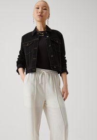 Veste en denim noire avec poches poitrine et surpiqûres dorées, associée à un pantalon blanc léger et ample avec une taille élastique.