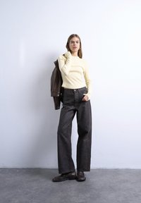 Νεαρή γυναίκα με κρεμ μακρυμάνικη μπλούζα, ψηλόμεσο φαρδύ σκούρο παντελόνι, καφέ παπούτσια, κρατώντας καφέ σακάκι στο ώμο, δίπλα σε λευκό τοίχο.