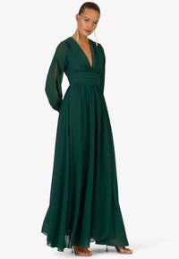 Langes grünes Kleid aus fließendem Stoff mit tiefem V-Ausschnitt, geraffter Taille und langen halbtransparenten Ärmeln. Verfügt über Schlitze an den Manschetten und einen subtilen Glanz.