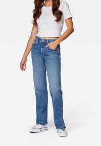 Mujer vestida con una camiseta corta blanca, jeans de tiro alto azul con dobladillos sin acabar y zapatillas blancas de plataforma, de pie con una mano en el bolsillo.
