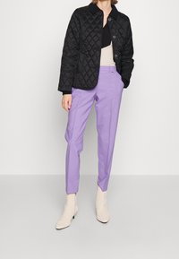 Czarna pikowana kurtka z okrągłym kołnierzem, zestawiona z jasnofioletowymi spodniami i beżowymi teksturowanymi botkami za kostkę.
