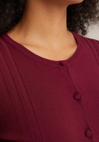 Bordo pletený top s kulatým výstřihem, vertikálním žebrováním a dvěma předními knoflíky na zapínání. Měkký materiál a přiléhavý střih.