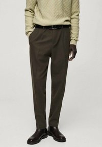 Pantalon marron sur mesure en tissu, avec une texture lisse, des plis à l'avant, des poches latérales, porté avec une ceinture noire et des bottes foncées.
