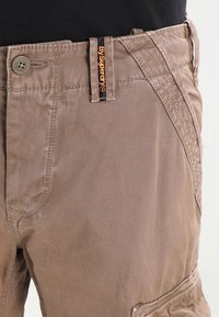 Los pantalones cortos cargo de algodón marrón presentan una textura suave, un cierre de botón y un distintivo detalle de costura a lo largo de los lados con una cinturilla de marca.