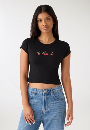 Czarny krótki t-shirt wykonany z miękkiej tkaniny, zdobiony trzema czerwonymi haftowanymi wiśniami na przodzie, zestawiony z jasnoniebieskimi dżinsami.