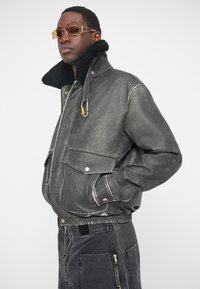 Veste en denim noir avec col en shearling, poches avant et coutures visibles. Le mannequin porte des lunettes teintées. Fini texturé et décoloré.