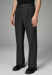 Pantalon noir à motif chevron, coupe droite, et une ceinture avec une boucle en métal. Porté avec des chaussures noires, mettant en valeur la texture et le design.