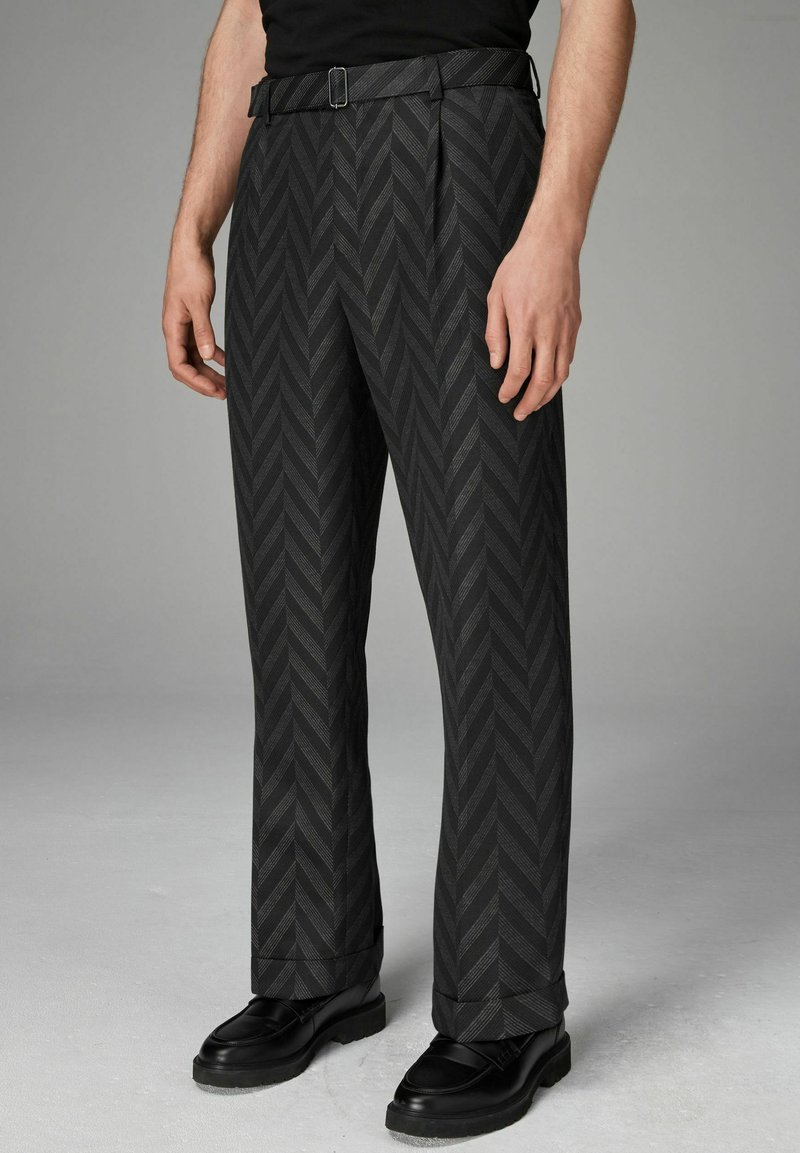 Pantalon noir à motif chevron, coupe droite, et une ceinture avec une boucle en métal. Porté avec des chaussures noires, mettant en valeur la texture et le design.