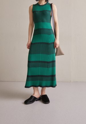 Femme portant une robe midi sans manches rayée verte et noire, des chaussures noires slip-on et un sac à main beige porté à l'épaule.