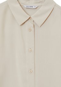 Camicetta beige con colletto, dotata di una chiusura a bottoni sulla parte anteriore e un'etichetta "CALLIOPE" di taglia S all'interno del colletto.