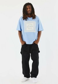Camiseta de gran tamaño en azul claro con un estampado gráfico en blanco que presenta las palabras "Project X Paris". Combinada con pantalones cargo negros holgados y zapatillas blancas.