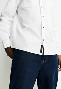 Hvid skjorte med knaplukning lavet af struktureret stof, med en brystlomme og grå knapper, parret med mørkeblå denim jeans.