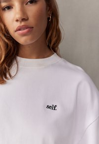 Bijela majica dugih rukava s zaobljenim izrezom, na prsima s crnim izvezenim tekstom "self.". Glatki, mekani pamučni materijal.