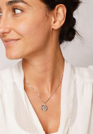 Silver necklace with a round pendant featuring a floral design, worn over a white blouse with vertical stripes.