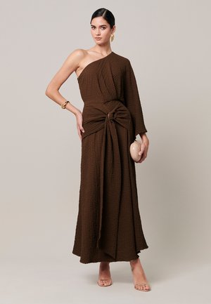 Woman in a textured brown one-shoulder dress with a knot detail, holding a beige clutch, wearing gold earrings and bracelet, standing against plain background.