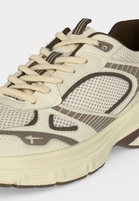 Chaussure de sport beige clair et marron avec des panneaux en mesh, semelle épaisse et amortissante, et lacets blancs attachés en nœud.