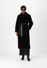 Abrigo largo de pelaje sintético negro con un amplio cuello, cintura ceñida y bolsillos laterales. Lleva encima de un vestido negro texturizado y está combinado con botas negras.